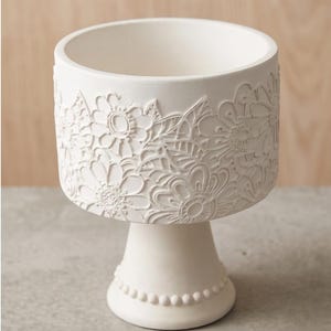 Könnte beinhalten: Eine matte, weiße Keramikschale auf einem Fuß mit einem erhabenen, geprägten Blumenmuster an der Außenseite des Beckens. Der Fuß der Schale ist mit einer einzelnen Reihe kleiner, gleichmäßiger Perlen am unteren Rand verziert.