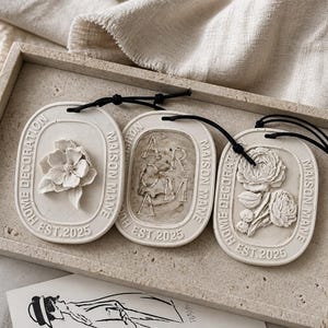 May include: Three cream-coloured oval home fragrance diffusers with black cords. Each has a raised floral design or the word "AROMA". The text "HOME DECORATION", "MAISON MANE", and "EST. 2025" are also visible on each.