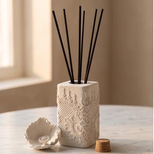 Puede incluir: Un difusor de varillas rectangular de color blanco con un patrón de encaje en relieve en el exterior. Seis varillas negras finas están insertadas en la abertura superior. Un adorno de flor de cerámica blanca y un tapón de corcho están al lado.