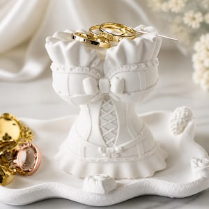 May include: A white matte corset-shaped jewellery holder with a ruffled top and base, featuring a central bow and lace-up detail. The piece sits on a matching scalloped tray, with two gold-toned rings resting on the top and several more on the tray.