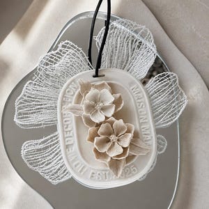 May include: An oval-shaped white ceramic hanging ornament featuring two sculpted beige flowers in the centre. The border is embossed with the text HOME DECORATION MAISON MAN EST 2025. A black cord is attached, and white mesh fabric wings extend from the sides.