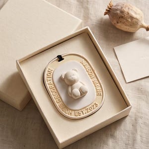 May include: An oval-shaped cream-colored ceramic ornament featuring a raised teddy bear figure in the center. The border is embossed with the text "MAISON MANE" and "HOME DECORATION EST. 2023". It includes a black cord for hanging and is presented in a box.