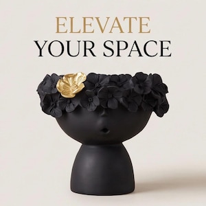 May include: A matte black planter shaped like a head with a small circular mouth opening. The top is adorned with a crown of sculpted black flowers, featuring one single metallic gold flower. Above the planter, the text reads ELEVATE YOUR SPACE.