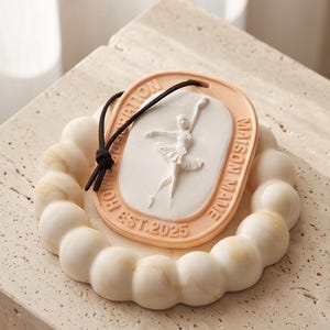 Op de afbeelding: Een ronde, crèmekleurige decoratieve plaquette met een textuur van bubbels langs de rand. Het midden toont een ovaal reliëf van een ballerina in een danshouding. De rand bevat de tekst "MAISON MANE" en "EST. 2025" met een zwart koord.