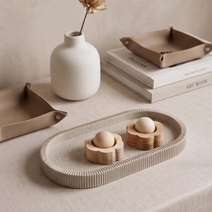 May include: An oval, ribbed, light-coloured tray holds two wooden flower-shaped egg cups, each containing a smooth, round wooden sphere. Nearby are two beige leather valet trays, a matte white vase with a dried flower, and two stacked books.
