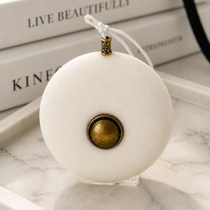 Puede incluir: Adorno redondo y blanco con un centro color latón y una parte superior decorativa. El adorno cuelga de una cuerda blanca y se exhibe en una bandeja de mármol. El fondo incluye libros blancos con el texto "LIVE BEAUTIFULLY" y "KINF".