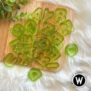 May include: A set of green plastic alphabet cookie cutters on a wooden surface. The cutters are in various shapes and sizes, and some are overlapping. The cutters are arranged in a random pattern.