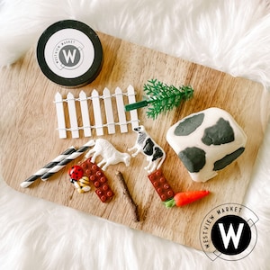 Peut inclure: Une planche en bois avec une clôture en bois blanc, une pâte à modeler en forme de vache, un petit arbre vert, une figurine de vache noire et blanche, une figurine d'agneau brun et blanc, deux figurines de coccinelles rouges, une paille rayée noire et blanche, deux blocs de construction en plastique rouge, un bâton brun et deux contenants avec le texte "Westview Market" et la lettre "W" en blanc.