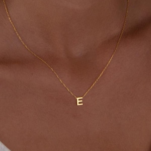 May include: A delicate gold-colored necklace featuring the letter "E" pendant. The pendant is small and centered on the chain. The necklace is worn around the neck, resting on a light-colored top.
