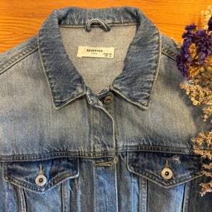 May include: A light blue denim jacket with a button closure and two front pockets. The jacket has a label that reads "Reserved" with the size "XXL" and "EUR 40" printed on it.
