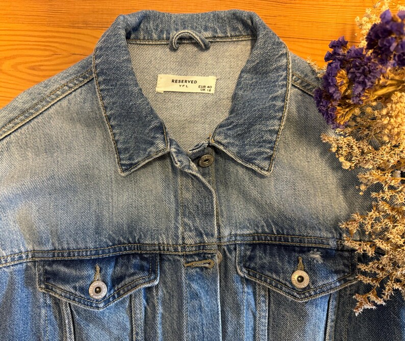 May include: A light blue denim jacket with a button closure and two front pockets. The jacket has a label that reads "Reserved" with the size "XXL" and "EUR 40" printed on it.