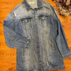 May include: A long, light blue denim jacket with distressed details and a button closure. The jacket has a classic collar and two front pockets.