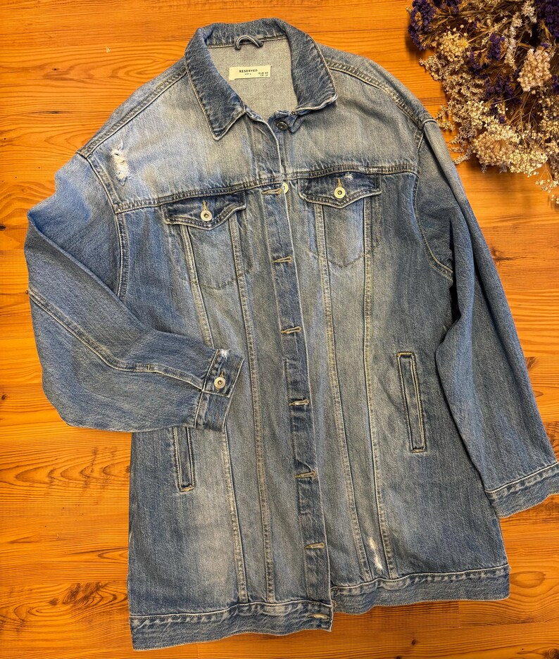 May include: A long, light blue denim jacket with distressed details and a button closure. The jacket has a classic collar and two front pockets.