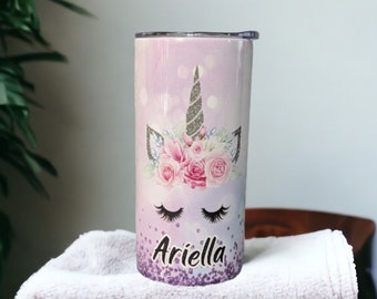 Könnte beinhalten: Ein Edelstahlbecher mit einem lila, rosa und weißen Einhorn-Design. Das Einhorn hat ein silbernes Horn und eine Blumenkrone. Der Becher ist mit dem Namen "Ariella" in silbernem Glitzer verziert.