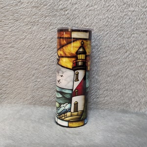 May include: A stainless steel tumbler with a colorful stained glass design of a lighthouse, waves, and birds. The lighthouse is white with red and black accents. The background is a sunset with orange, yellow, and blue colors.
