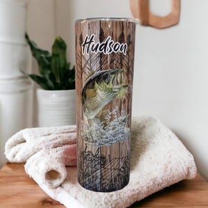 May include: A stainless steel tumbler with a wood grain design and a largemouth bass jumping out of the water. The tumbler has the name "Hudson" printed on it.