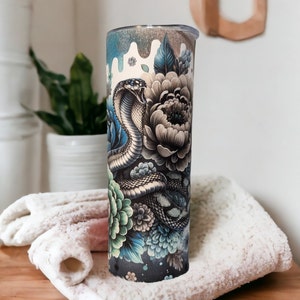 May include: A cylindrical tumbler featuring a detailed graphic print of a coiled cobra with an open mouth, surrounded by large, layered floral blossoms in shades of teal, blue, and grey, with a dripping paint effect at the top rim.