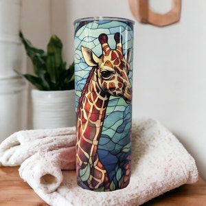 May include: A stainless steel tumbler with a colorful stained glass design featuring a giraffe's head. The giraffe has a brown and yellow patterned coat and a blue background.