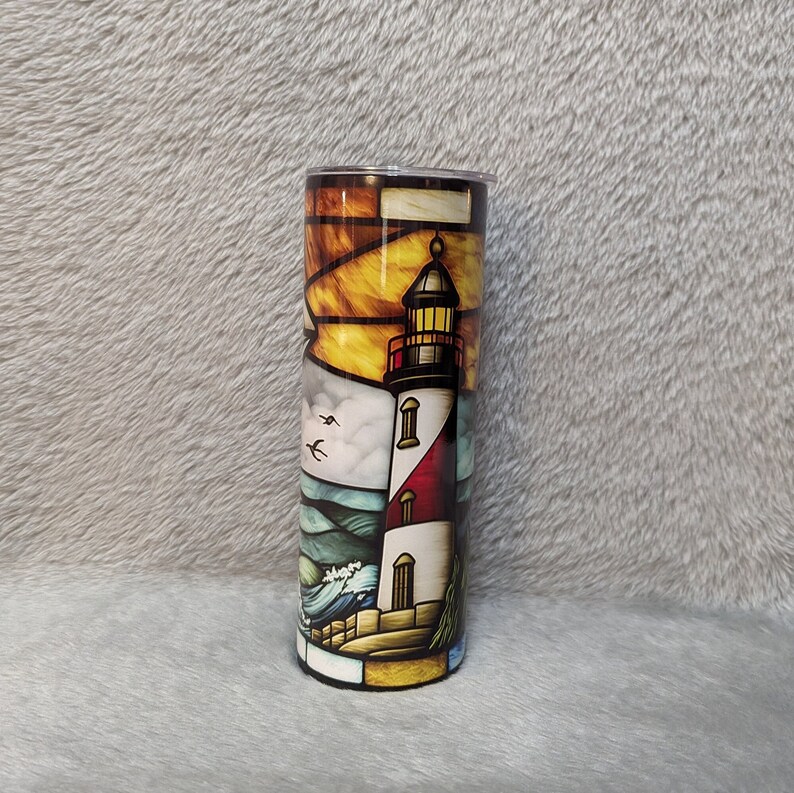 May include: A stainless steel tumbler with a colorful stained glass design of a lighthouse, waves, and birds. The lighthouse is white with red and black accents. The background is a sunset with orange, yellow, and blue colors.