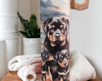 May include: A stainless steel tumbler with a colorful illustration of a Rottweiler dog and two puppies. The background features a watercolor-style sky and beach scene.