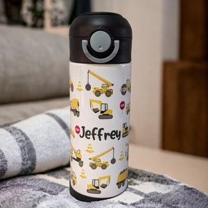 May include: A yellow and white insulated water bottle with a black lid and a flip top. The bottle has a construction theme with images of construction vehicles and the name "Jeffrey" printed on the side.
