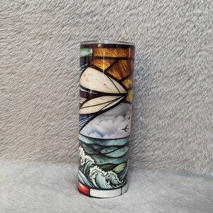 May include: A stainless steel tumbler with a stained glass design of a seascape. The design features a sunset, clouds, birds, and waves in shades of blue, green, yellow, and orange.