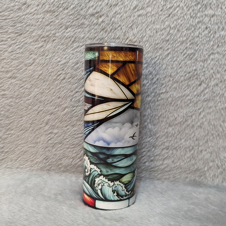 May include: A stainless steel tumbler with a stained glass design of a seascape. The design features a sunset, clouds, birds, and waves in shades of blue, green, yellow, and orange.