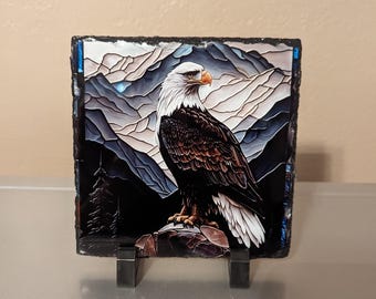 May include: A square, decorative slate tile featuring a majestic bald eagle perched on a rock. The eagle has white feathers on its head and a brown body. The background depicts a mountain range in shades of gray and blue. The tile is displayed on a metal stand.