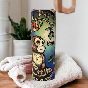 May include: A stainless steel tumbler with a colorful design featuring a monkey, flowers, and vines. The tumbler has a blue, green, and yellow background with a moon and the name "Kathy" printed on it.