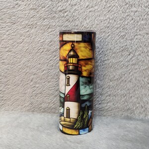 May include: A stainless steel tumbler with a colorful stained glass design of a lighthouse. The lighthouse is white with red and black accents. The background is a blue and yellow sky with waves crashing against the shore.