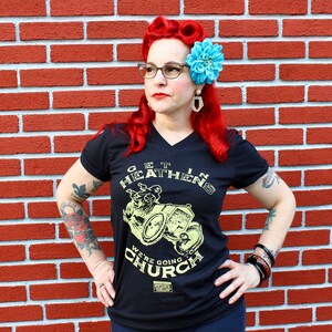 May include: A woman with red hair and a blue flower in her hair is wearing a black t-shirt with a yellow graphic of a car and the text "Get in Heathens We're Going to Church".