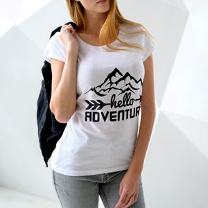 Puede incluir: Camiseta blanca con un gr&aacute;fico de monta&ntilde;as negras y el texto "hello adventure" con una flecha apuntando hacia la derecha.