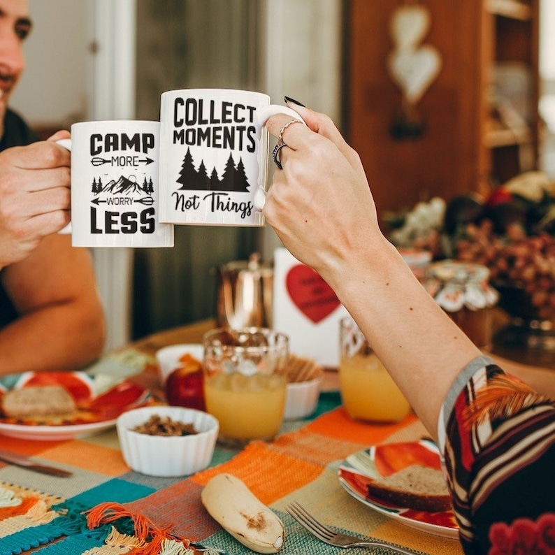 Puede incluir: Dos tazas de cer&aacute;mica blanca con texto negro y gr&aacute;ficos. La taza de la izquierda dice "Camp More Worry Less" con un gr&aacute;fico de monta&ntilde;a y flecha. La taza de la derecha dice "Collect Moments Not Things" con un gr&aacute;fico de bosque.