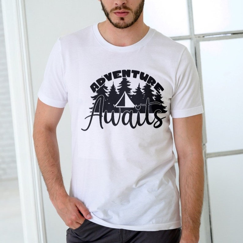 Puede incluir: Camiseta blanca con un dise&ntilde;o gr&aacute;fico negro que dice "Adventure Awaits" con una silueta de una tienda de campa&ntilde;a y &aacute;rboles.