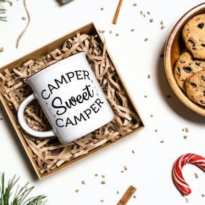 Puede incluir: Taza de cer&aacute;mica blanca con texto negro que dice "Camper, Sweet Camper" en una fuente decorativa. La taza est&aacute; en una caja de regalo de papel marr&oacute;n llena de papel triturado.