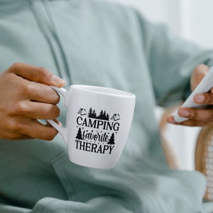 Puede incluir: Taza de cer&aacute;mica blanca con texto negro que dice "Camping favorite therapy" y un gr&aacute;fico de pinos y tiendas de campa&ntilde;a.