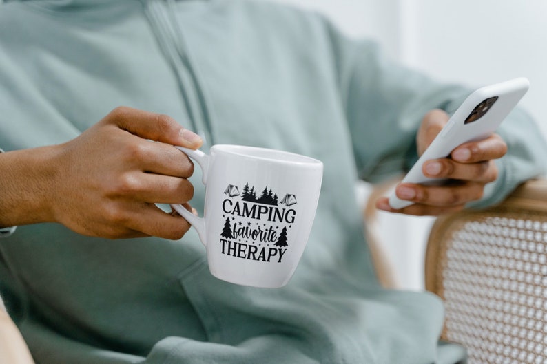 Puede incluir: Taza de cer&aacute;mica blanca con texto negro que dice "Camping favorite therapy" y un gr&aacute;fico de pinos y tiendas de campa&ntilde;a.