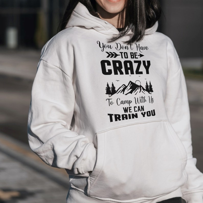 Puede incluir: Sudadera blanca con texto negro que dice "You Don't Have To Be Crazy To Camp With Us We Can Train You". El texto est&aacute; rodeado por un gr&aacute;fico de monta&ntilde;a y pinos.