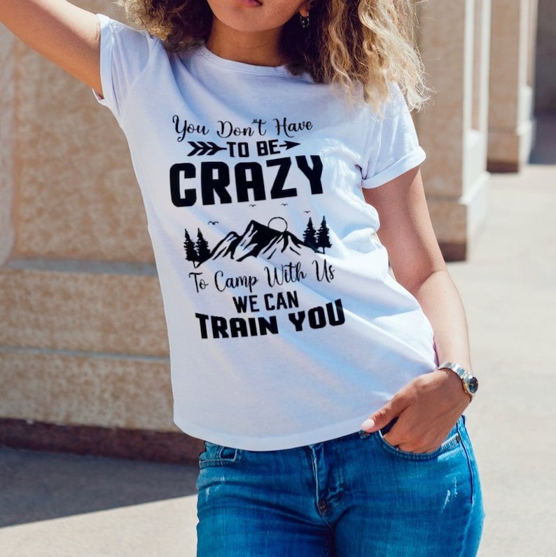 Puede incluir: Camiseta blanca con texto negro que dice "You Don't Have To Be Crazy To Camp With Us We Can Train You". El texto est&aacute; rodeado de una escena de monta&ntilde;a con &aacute;rboles y un sol.