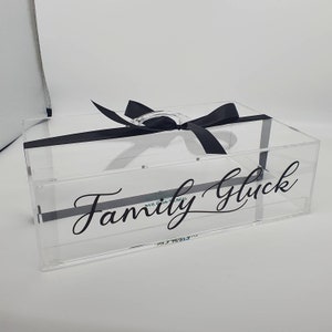 May include: A clear acrylic box with the words "Family Glück" in black script. The box is tied with a black satin ribbon and has a clear handle. The box is rectangular and sits on a white surface.