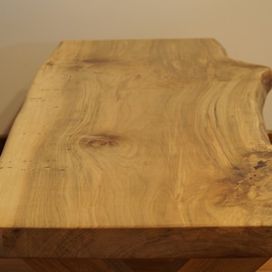 May include: A large, natural wood coffee table with a live edge top and a knot in the wood. The table has a light brown stain and a natural wood finish.