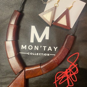 May include: A wooden necklace with a triangular pendant that says "420". The necklace is made of dark brown wood and has a simple design. The necklace is accompanied by a pair of wooden earrings with a white and brown triangular design. The earrings are attached to a silver chain.