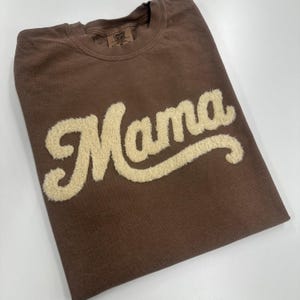 May include: A brown t-shirt with the word "Mama" in a cream-colored, textured font. The shirt is folded neatly, and a small tag is visible near the collar. The overall aesthetic is casual and comfortable.