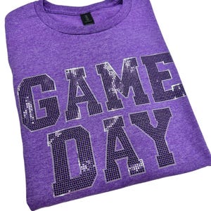 May include: A heathered purple t-shirt featuring the text "GAME DAY" in large, block-style letters. The lettering is composed of numerous small, reflective silver sequins that create a textured, metallic appearance against the fabric.