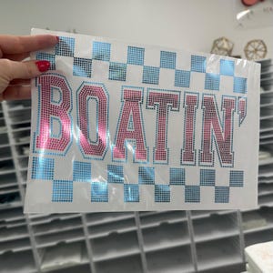 May include: A sheet of iron-on rhinestones with the word "BOATIN'" in pink and red rhinestones. The design includes a blue and black checkered pattern. The sheet is held up by a hand.