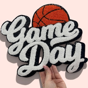 May include: A tufted wall hanging featuring the text Game Day in white script lettering with a black outline. An orange basketball with black lines is positioned above the word Game. The entire piece has a textured, plush appearance.
