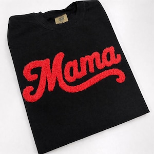 May include: Black crew neck t-shirt with the red word "Mama" in a bold, textured font. The shirt has a tag at the neckline. The design is centered on the front of the shirt.