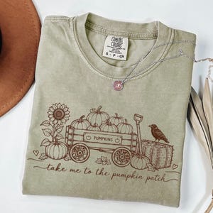 May include: A sage green short-sleeve t-shirt featuring a brown graphic print of a wooden wagon filled with pumpkins, a sunflower, a crow on a hay bale, and the text "take me to the pumpkin patch". The shirt has a Comfort Colors brand label.