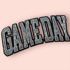 May include: A decorative patch featuring the word GAMEDAY in block letters. The letters are covered in iridescent silver sequins and outlined with a thick black border that has a glittery texture, creating a bold and high-contrast appearance.