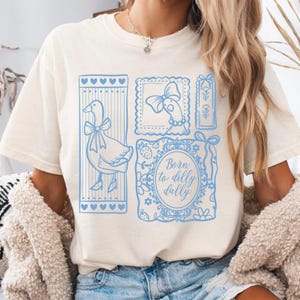 May include: Cream-colored t-shirt featuring a light blue graphic design. The design includes a goose with a bow, framed illustrations, and the text "Born to dilly dally". The shirt has short sleeves and a relaxed fit.
